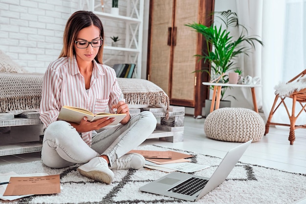 Female student sitting on floor watching webinar on laptop and taking notes in textbook Copy space