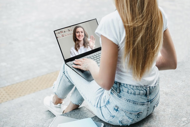 Studentessa seduta con il laptop sulle scale e fare una videochiamata alla sua amica