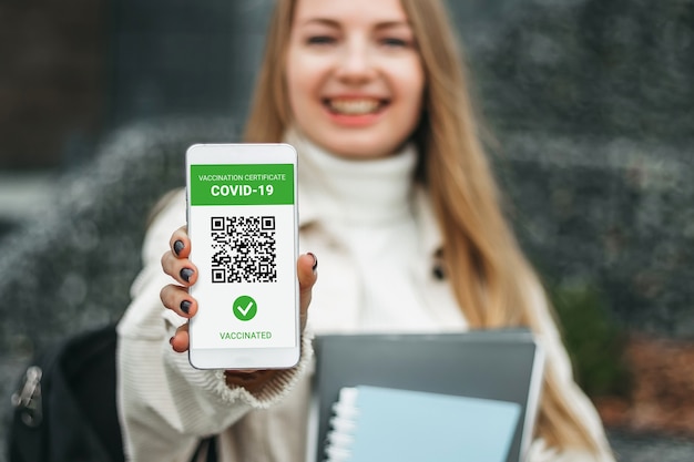 Female student smiling and showing a mobile phone screen with an application about vaccination against coronavirus, covid-19 against the background of a university
