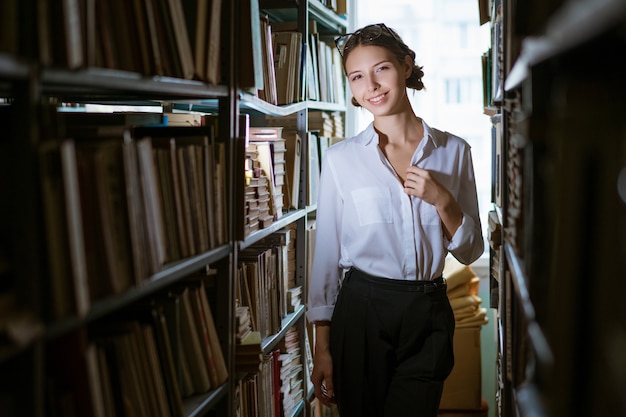 female student in  white shirt stands between the rows in the library, bookshelves worth of books. Dark photo