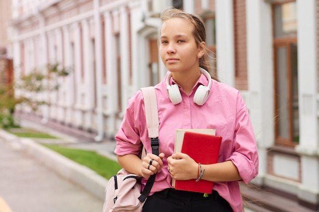 Female student with a backpack and books with headphones outdoors on a college campus