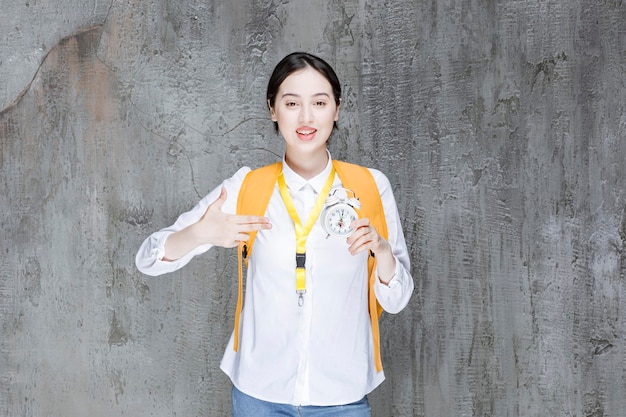 Female student with yellow backpack holding clock. High quality photo