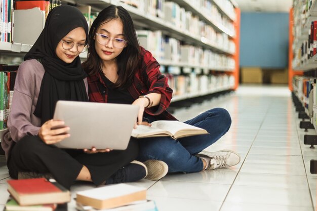 female students finishing assignment together using laptop and book at the library