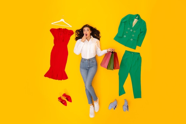 Female stylist holding shopper bags lying among clothes yellow background