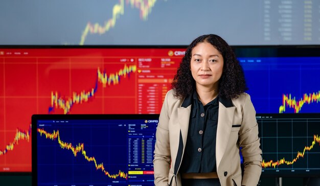 Female successful professional broker trader investor stand smile put hands in pants pocket look at camera in front computer monitor screen with stock and bitcoin cryptocurrency graph chart analysis.