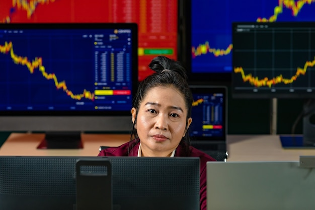 Female successful professional trader investor sit smile look at camera surrounded by computer monitor screen with graph chart stock and bitcoin cryptocurrency analysis growth report in trading room.