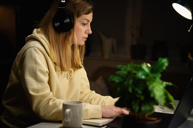 自宅でラップトップのオンライン教育を使用して夜の部屋で勉強しているヘッドフォンを身に着けている女性の学生