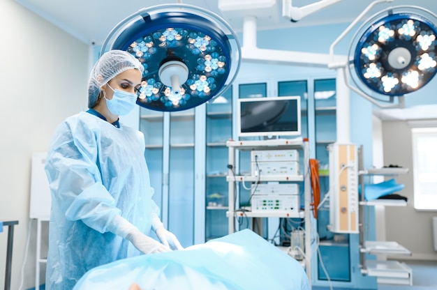 Female surgeon in gown and mask in operating room, lamp on background operation