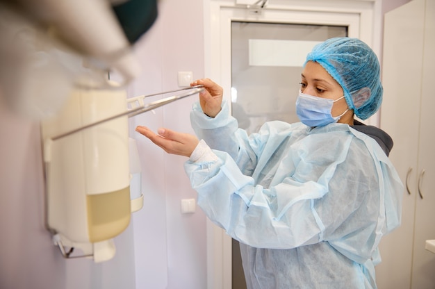 Female surgeon in medical protective mask and professional
workwear standing in washing room of surgery clinic and using
dispenser sanitizer for disinfecting and sanitizing hands before
surgery