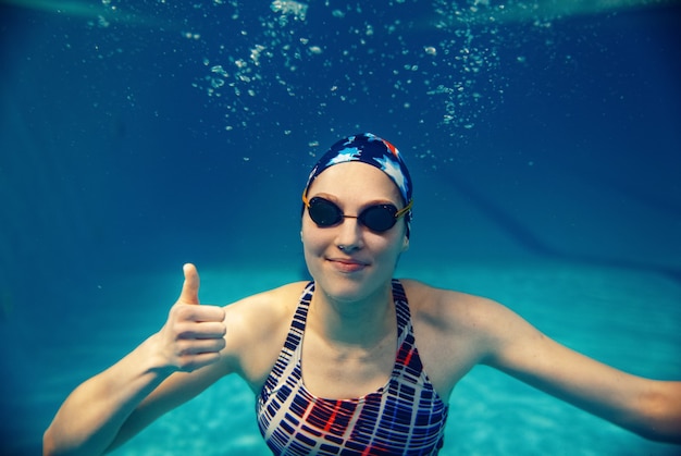 수영복, 수영 모자 및 안경 여성 수영 선수가 수영장에서 수중 엄지 손가락을 보여줍니다.
