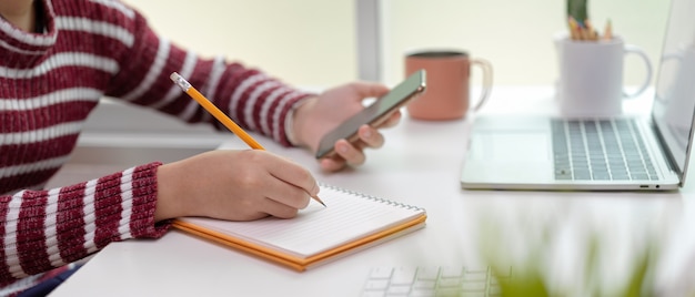 Female  taking note on blank notebook while using smartphone on office desk