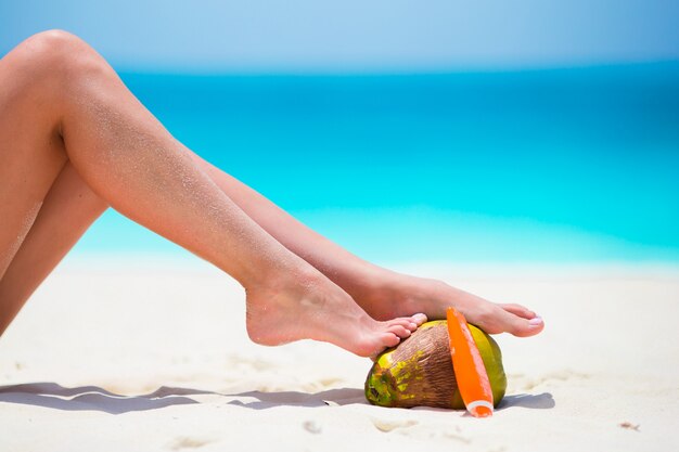 Female tanned smooth legs with suncream and coconut on white beach