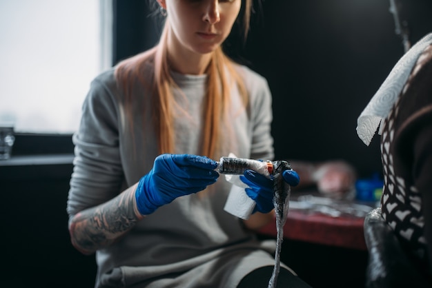 Female tattooist in blue sterile gloves prepares tattoo machine, master in salon. Professional tattooing in studio