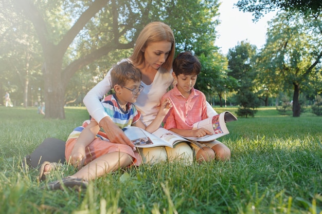 Female teacher reading with her little students, sitting in the grass at public park