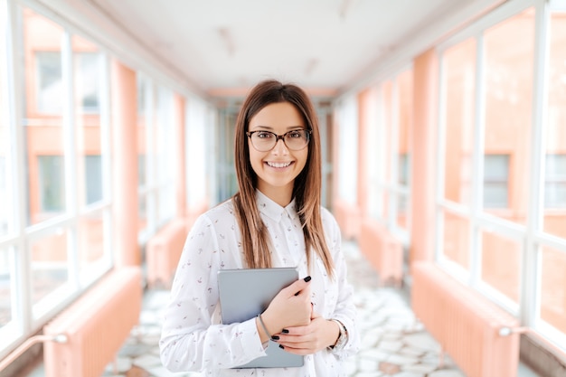 female teacher with eyeglasses and brown hair holding tablet in arms while standing in the hall