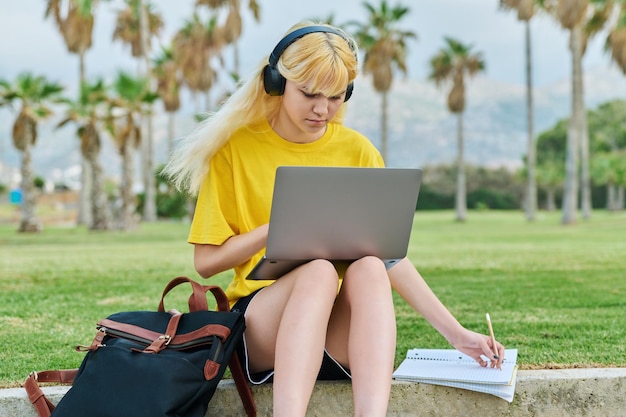 Female teenage student in headphones sitting in park with laptop