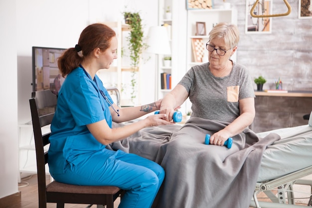 Female therapist helping senior woman in nursing home do to exercises with dumbbells.