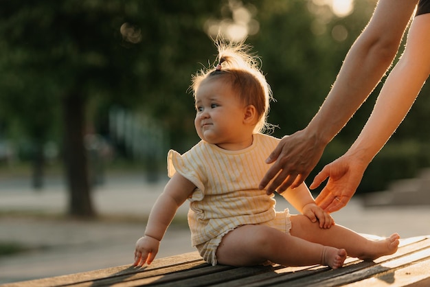 A female toddler at the bench near a hand of a mom in the park in the sun lights