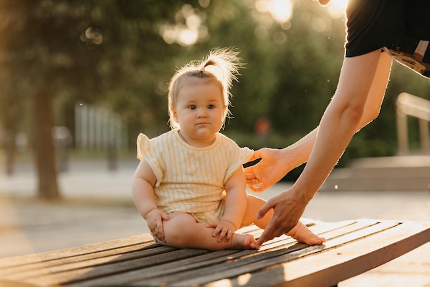 A female toddler at the bench near a hand of a mom in the park in the sun lights