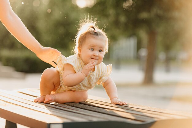 A female toddler at the bench near a hand of a mom in the park in the sun lights