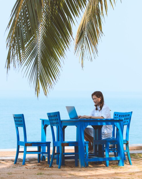 Female tourism sitting at blue desk beside sand beach with leaves of coconut tree and using laptop computer for online working.