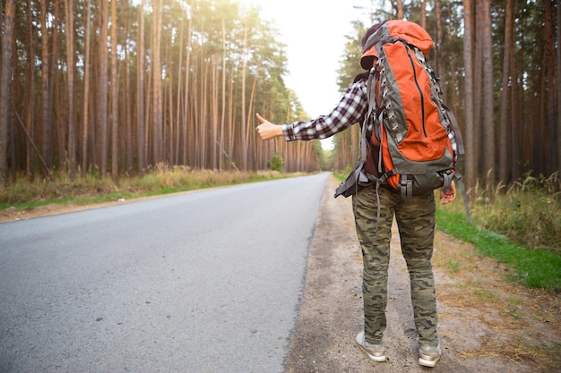 Фото Туристка в клетчатой рубашке с оранжевым большим рюкзаком возле шоссе в лесу голосует за то, чтобы ее подвезли. автостоп, внутренний туризм. путешественник, одиночное приключение, поездка, поход