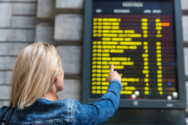 女性観光客は駅の時刻表を見て、ヨーロッパを旅行します。ヨーロッパの鉄道による輸送、情報ディスプレイでの乗客、快適な観光と旅行