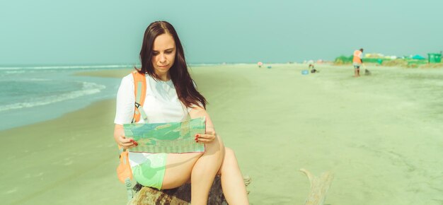 Female tourist reading map near sea barefoot woman with\
backpack sitting on driftwood and examining map while resting on\
sandy beach near waving sea