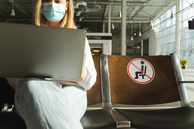 Female tourist sitting in airport lobby chair with social distancing sign and using laptop