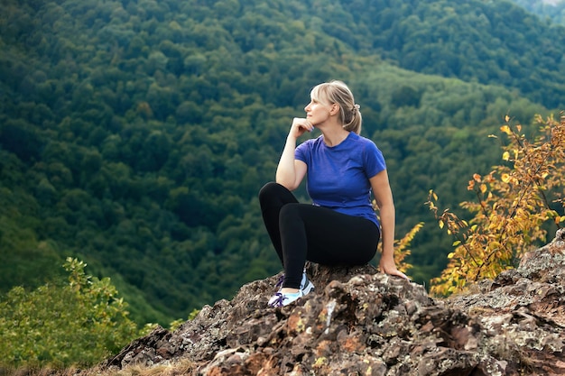 Female tourist in sportswear sits on a high mountain, rests and enjoys the view of nature