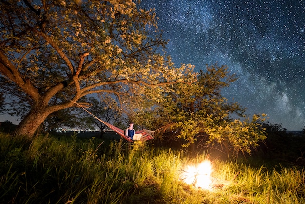 Turista femminile che utilizza il suo computer portatile nel campeggio alla notte. donna seduta sull'amaca vicino al fuoco sotto gli alberi e il bel cielo notturno pieno di stelle e via lattea
