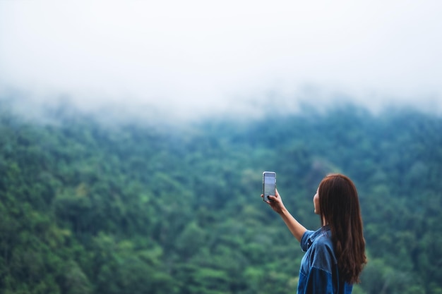 A female tourist using smartphone to take a photo of a beautiful foggy mountains in the forest