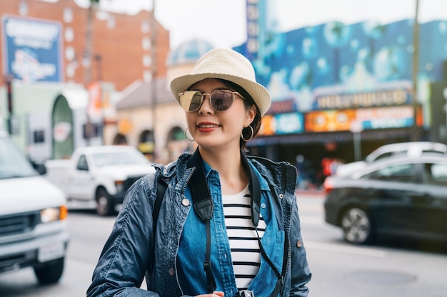 female tourist walking on the street visiting the city. Asian woman smiling in hat and sunglasses. elegant traveler enjoy the downtown lifestyle.