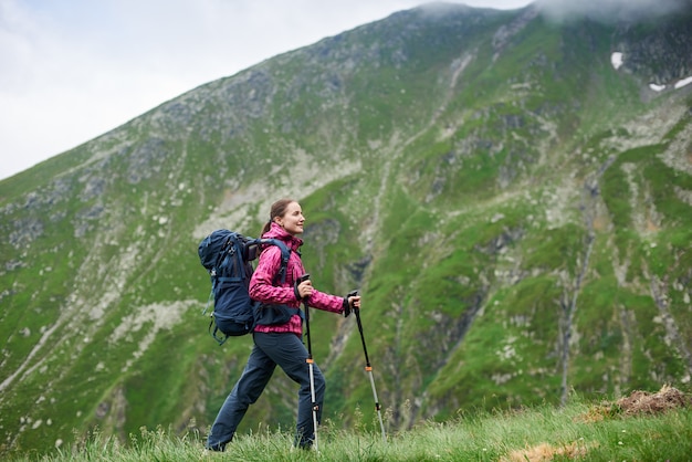 Female tourist walking up green grassy slope with walking stick and backpack in front of beautiful rocky mountains