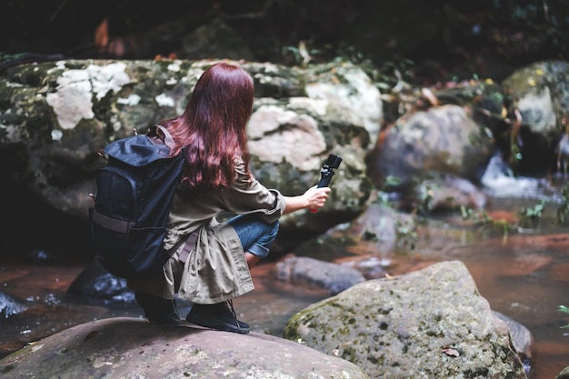 배낭을 메고 숲속의 폭포를 액션 카메라로 촬영하는 여성 관광객