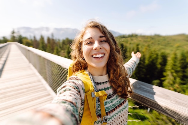 Photo female tourist with hiking backpack takes selfie with phone on hiking bridge in forest environment