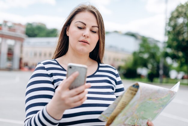 Female tourist with smartphone and paper map in hands looking for direction outdoors