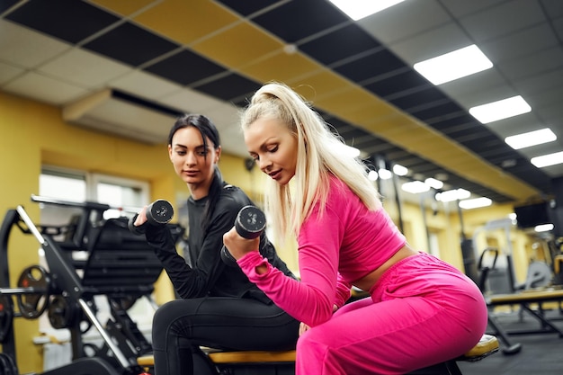 Female trainer shows the client exercise for biceps with dumbbell while sitting on the bench in gym