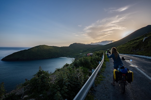 羊の写真とアイルランドのアキル島のキーム湾のパノラマビューを撮る女性旅行者サイクリスト