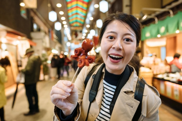 꼬챙이 음식을 들고 카메라를 마주하는 여성 여행자는 일본에서 이국적인 간식을 먹는 것에 흥분합니다. 카메라를 보고 있는 아시아 유튜버가 팬들에게 타코 타마고라는 이상한 일본 음식을 보여주고 있습니다.