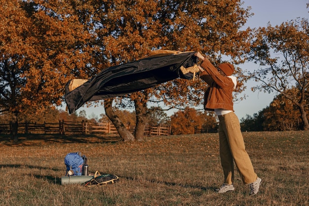 Female traveler pitching tent at campsite in autumn forest on sunny day