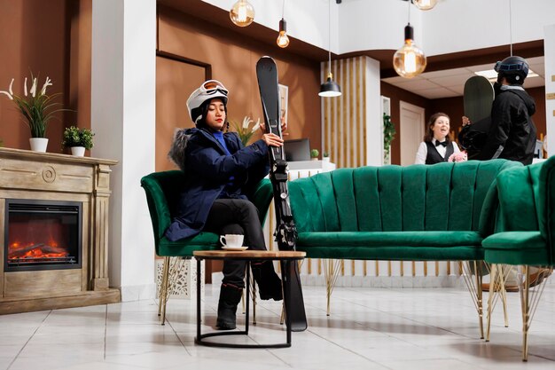 Female traveler seen in lounge area fully equipped for wintersports with snow gear and winter jacket Woman holding skiing skis seated on sofa while man with snowboard arrives at front desk