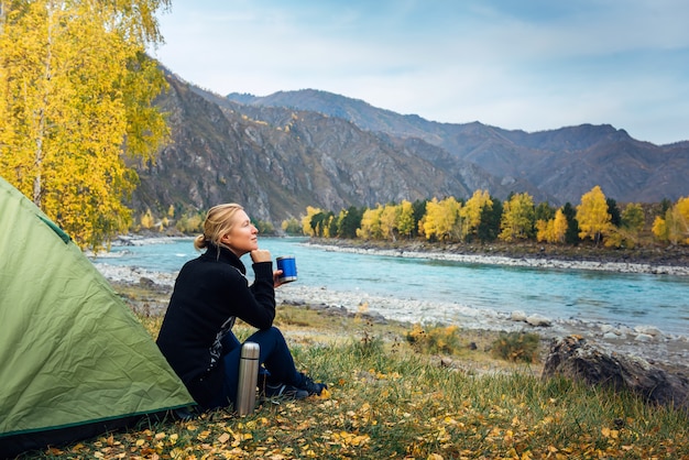 Female traveler sitting on grass near the tent, drinking coffee from thermos and admiring the beautiful view of river and mountains. Morning woman tourist, pleasure in the journey