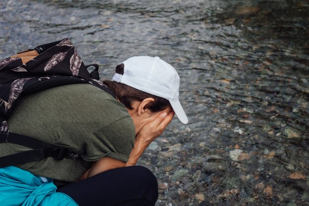 バックパックを背負った白い帽子をかぶった女性旅行者は、きれいな冷たい川の水で顔を洗います