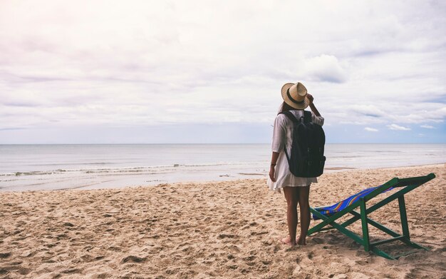 海沿いのビーチで一人で立っている帽子とバックパックを持つ女性旅行者