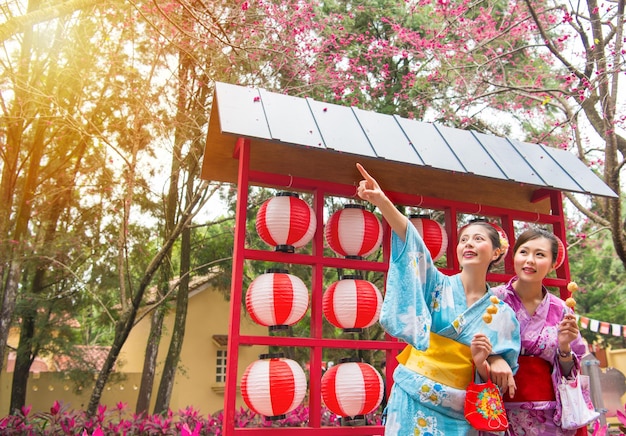 着物を着た女性旅行者が桜の花を指さし、おいしいつくねの串焼きを一緒に持ち、お祭りの提灯通りで美しい花を見ています。