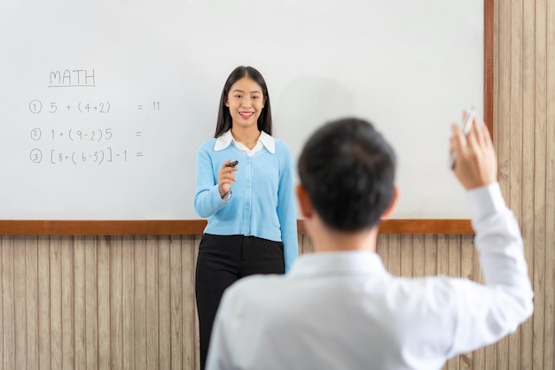 Female tutor standing in front of whiteboard is pointing to student for ask question and young students raising hands in the air to answer a question when learning in the classroom