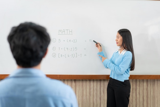 Foto tutor femminile in piedi davanti alla lavagna e che scrive equazioni matematiche a bordo per spiegare allo studente durante la lezione di matematica in classe