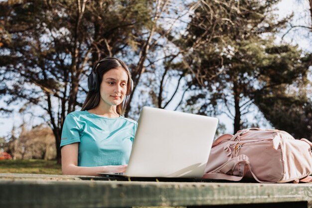 Female university student using a laptop in campus natural\
park. turquoise t shirt and pink backpack