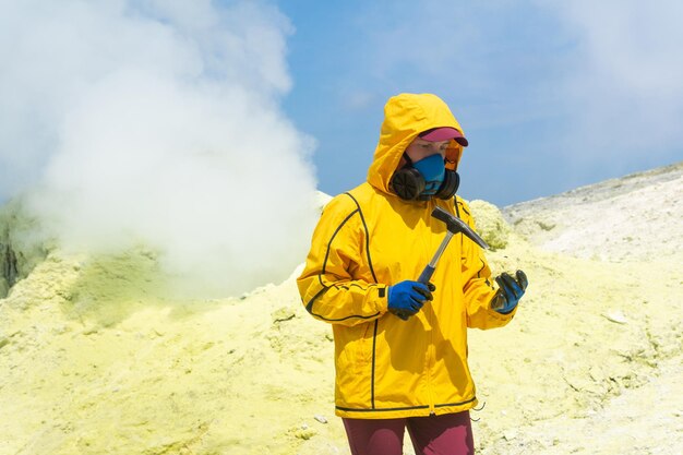 Female volcanologist on the background of a smoking fumarole examines a sample of a sulfur mineral with a geological hammer